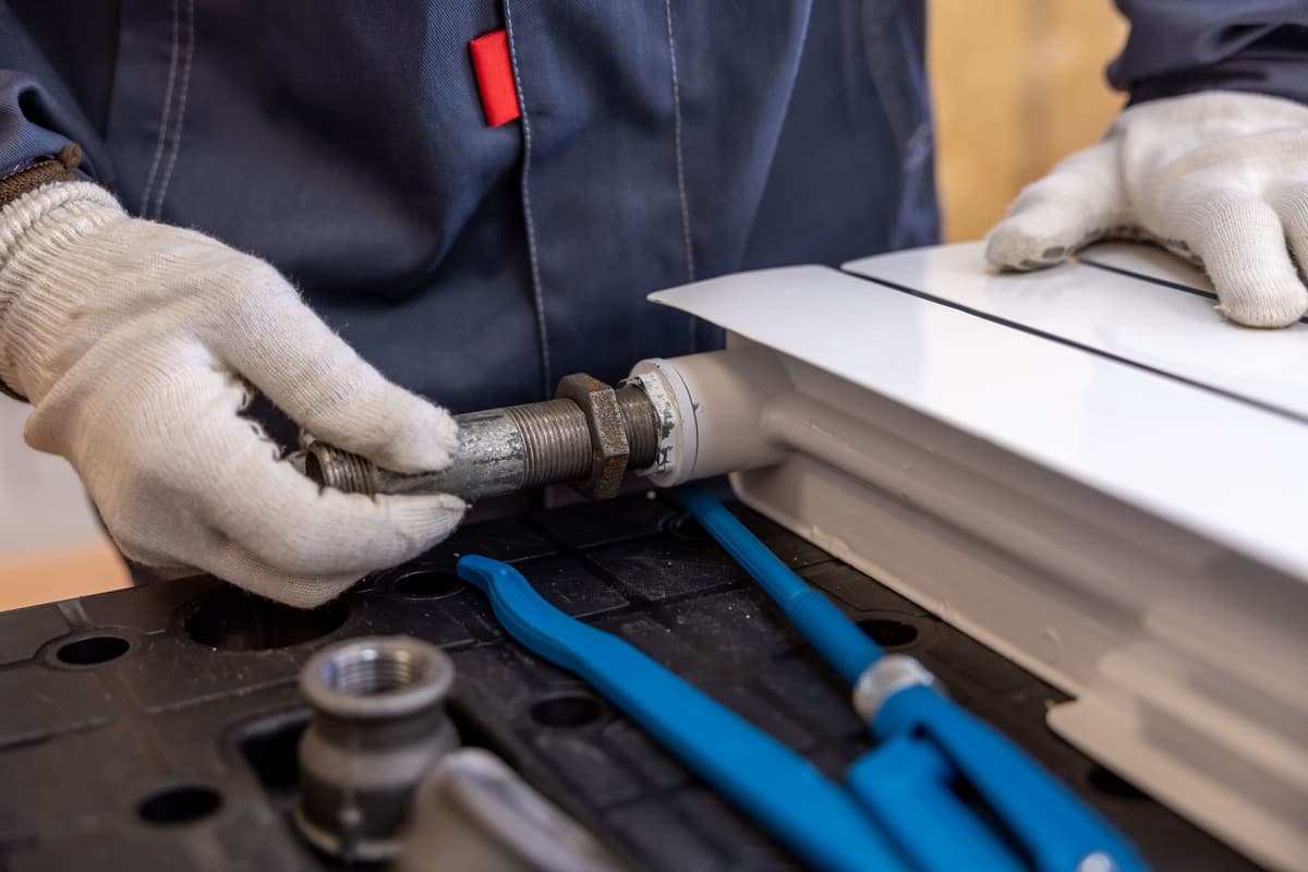 Gloved hands fitting a threaded pipe to a radiator, pipe wrenches on the bench